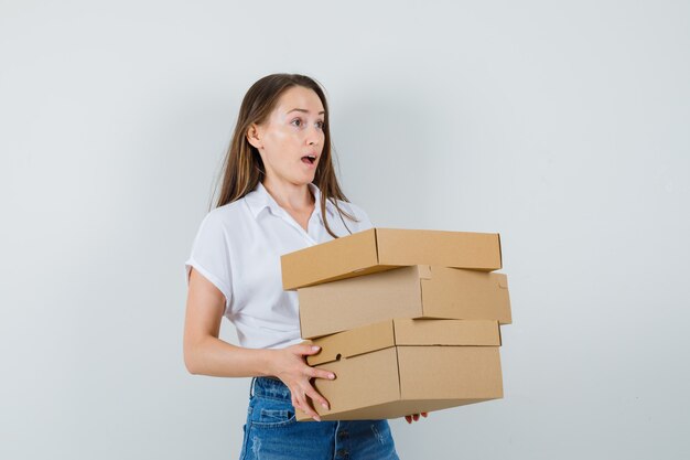 Bella dama llevando cajas en blusa blanca y mirando preocupado, vista frontal.