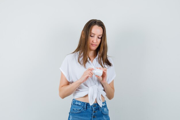 Bella dama mirando una botella de píldoras en blusa blanca, jeans y mirando atenta, vista frontal.