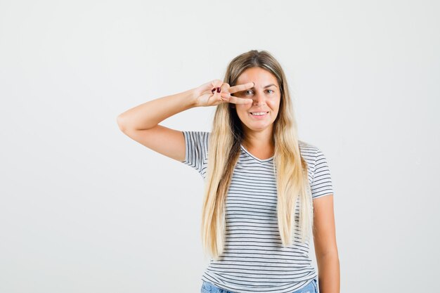 Bella dama mostrando el signo v sobre un ojo en camiseta y luciendo alegre. vista frontal.