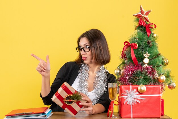 Bella dama de negocios en traje con gafas apuntando a algo sentado en una mesa con un árbol de Navidad en la oficina