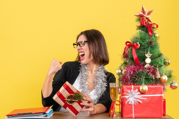 Bella dama de negocios en traje con gafas sosteniendo su regalo con orgullo sentado en una mesa con un árbol de Navidad en la oficina