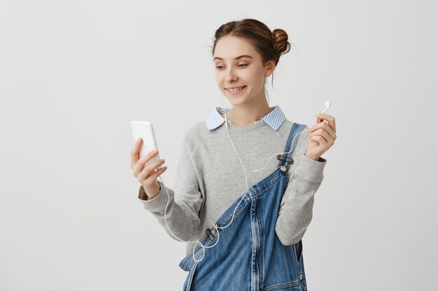 Bella dama en overol de mezclilla mirando en su teléfono sonriendo ampliamente. Esposa moderna y amigable que habla por Skype con su esposo mientras está fuera por negocios. Concepto de comunicación