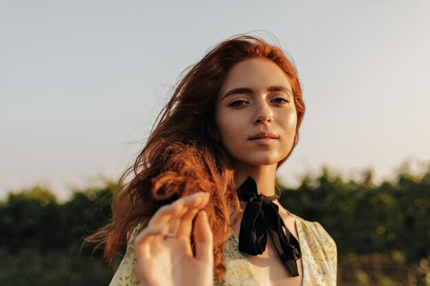 Bella dama con pelo ondulado foxy, lindas pecas y vendaje negro en el cuello en vestido encantador de verano mirando al frente al aire libre