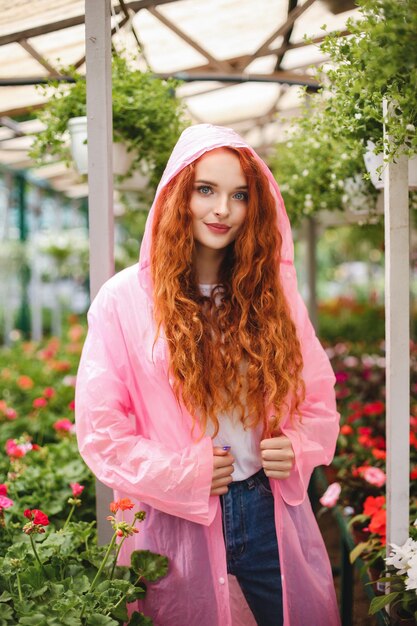 Bella dama con el pelo rizado pelirrojo de pie en un impermeable rosa y mirando soñadoramente a la cámara mientras pasa tiempo en el invernadero