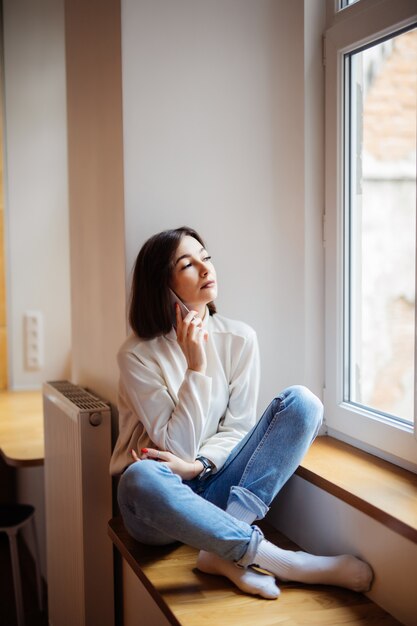 Bella dama en la sala sentada cerca de la ventana en ropa casual blue jeans