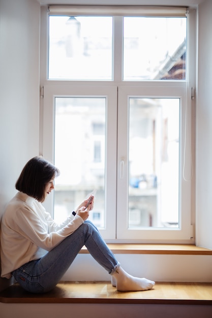 Foto gratuita bella dama en la sala sentada cerca de la ventana en ropa casual con teléfono