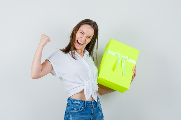 Bella dama sosteniendo la caja mientras muestra el gesto de ganador en blusa blanca, jeans y mirando alegre, vista frontal.