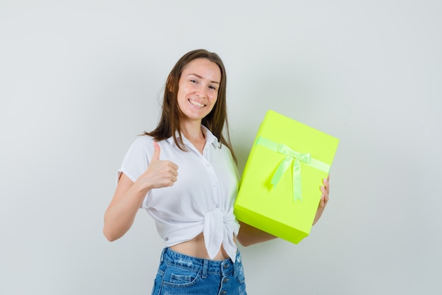 Bella dama sosteniendo la caja mientras muestra el pulgar hacia arriba en blusa blanca, jeans y luciendo positiva. vista frontal.