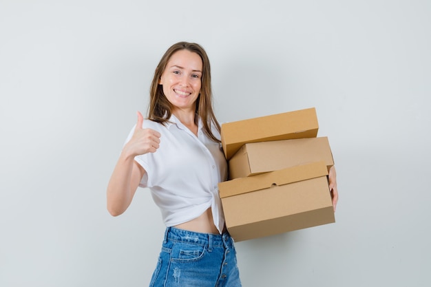 Bella dama sosteniendo cajas mientras muestra el pulgar hacia arriba en una blusa blanca y parece complacida, vista frontal.