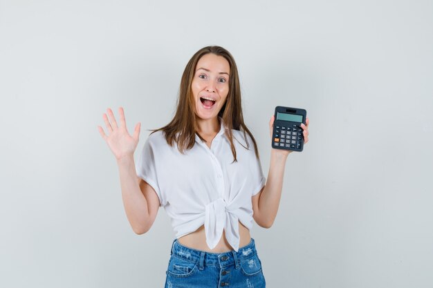 Bella dama sosteniendo la calculadora mientras agita la mano en blusa blanca, jeans y mirando enérgico, vista frontal.