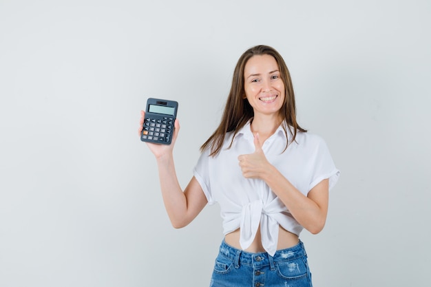 Bella dama sosteniendo la calculadora mientras muestra el pulgar hacia arriba en blusa blanca, vista frontal de jeans.