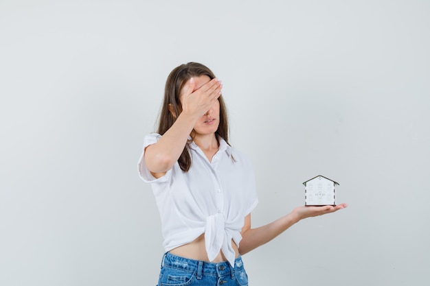 Bella dama sosteniendo la casa modelo mientras sostiene la mano en la cabeza con una blusa blanca y luce estresante, vista frontal.