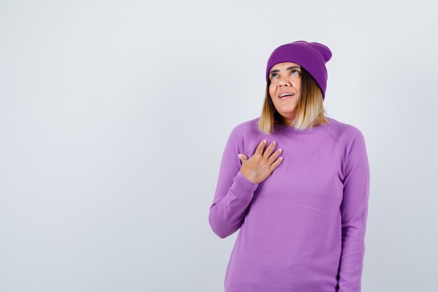 Bella dama en suéter, gorro manteniendo la mano en el pecho y mirando emocionada, vista frontal.