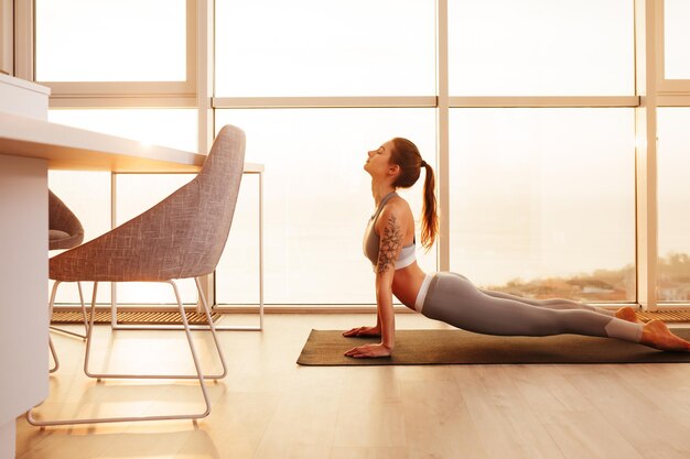 Bella dama en top deportivo y polainas practicando yoga en colchoneta de yoga en casa con grandes ventanas hermosas en el fondo