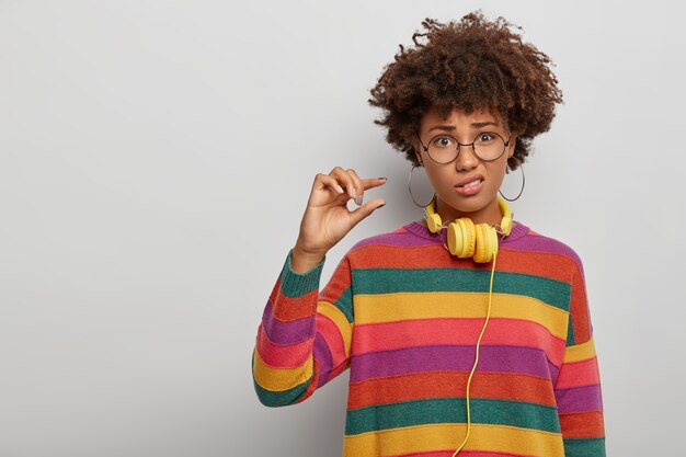 La bella dama triste muestra una cosa pequeña, hace un pequeño gesto, frunce el ceño con disgusto, se viste con ropa informal, usa grandes gafas redondas