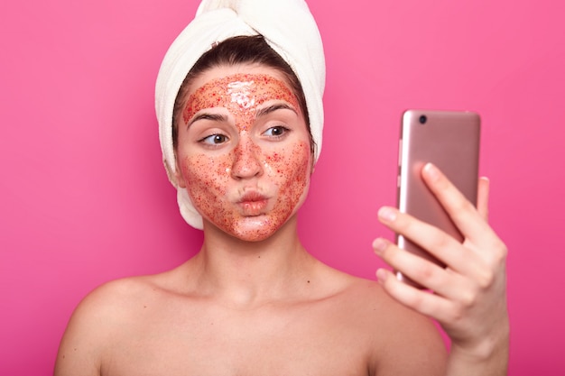 La bella modelo femenina tiene una máscara exfoliante en la cara, envuelta en una toalla, posa semidesnuda, toma selfie en el teléfono inteligente, aislada en rosa, se siente emocional, mantiene los labios redondeados. Concepto de cosmetología