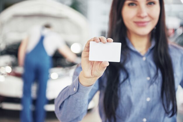 Una bella mujer guarda una tarjeta de visita del centro de servicio de automóviles. El mecánico inspecciona el auto debajo del capó en el fondo