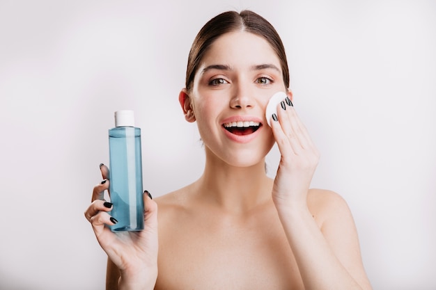 Bella mujer hidrata delicadamente la piel con tónico cosmético. Retrato de dama con piel sana sin maquillaje en pared aislada.