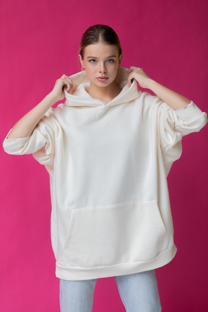 Bella mujer rubia con una sudadera blanca y vaqueros azules posando sobre un fondo rosa