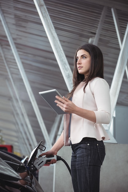 Bella mujer con tableta digital mientras carga el coche eléctrico