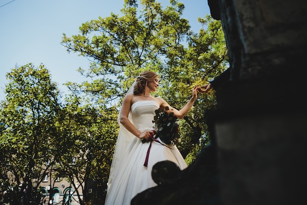 La bella novia mantiene un ramo de novia y de pie cerca del edificio