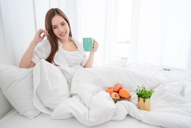 Belleza asiática mujer vestido blanco cabello largo mano sostener bebida caliente hora de la mañana en la cama blanca con almejas y alegre dormitorio blanco fondo interior