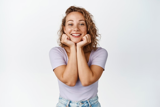 Belleza. Bonita mujer rubia con el pelo corto y rizado, cara magra en las manos y sonriendo, mirando a la cámara feliz, fondo blanco.