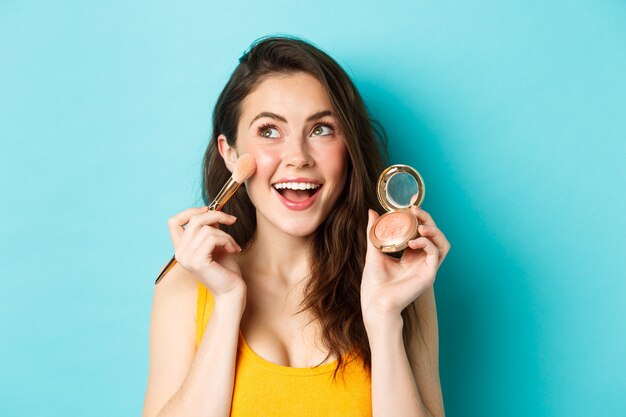 Belleza. Cerca de la hermosa mujer joven mira la esquina superior izquierda y sonríe mientras aplica rubores en las mejillas con pincel de maquillaje, de pie feliz contra el fondo azul.