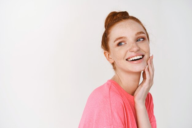 Belleza y cuidado de la piel Chica sonriente con el pelo de jengibre peinado en un moño desordenado tocando la piel perfecta y brillante y sonriendo mostrando la cara sin maquillaje de fondo blanco