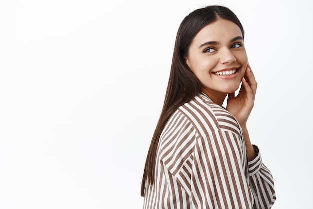 Belleza y cuidado de la piel La mujer caucásica moderna gira la cabeza detrás del hombro y mira a la izquierda en el espacio de la copia sonriendo complacido tocando la cara fondo blanco