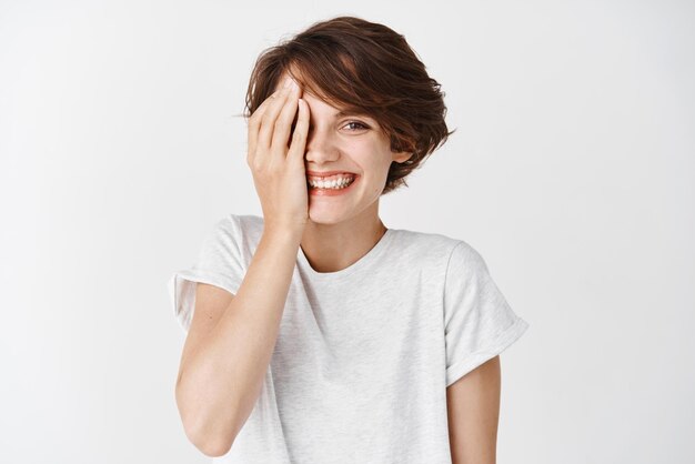 Belleza y cuidado de la piel Mujer joven alegre en camiseta que cubre la mitad de la cara y sonriendo feliz a la cámara de pie sobre fondo blanco.