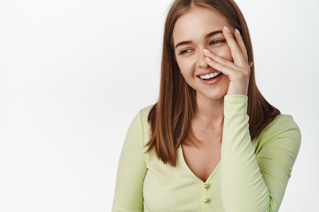 Belleza y cuidado de la piel. Mujer rubia tierna y hermosa riendo, sonrisa genuina, tocando una piel perfecta y brillante sin maquillaje, sonriendo y mirando a un lado, fondo blanco