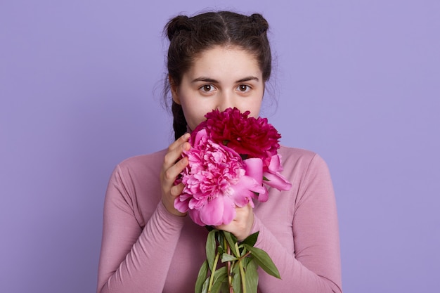 Belleza delicada mujer oliendo flores de primavera y con expresión encantadora, dama con coletas sosteniendo flores aisladas sobre pared lila.