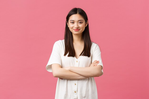 Belleza, emociones de la gente y concepto de estilo de vida de verano. Tierna joven asiática en lindo vestido sonriendo a cámara, de pie con las manos cruzadas y mirada profesional determinada, fondo rosa.