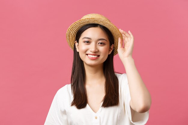 Belleza, emociones de la gente y concepto de ocio y vacaciones de verano. Chica asiática elegante y de moda con sombrero de paja y un lindo vestido blanco sonriendo a la cámara, disfrutando de las compras, fondo rosa.