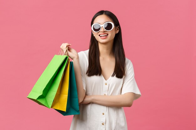 Belleza, emociones de la gente y concepto de ocio y vacaciones de verano. Chica asiática feliz despreocupada de vacaciones, turista con bolsas de la compra y gafas de sol, sonriendo satisfecho, fondo rosa.