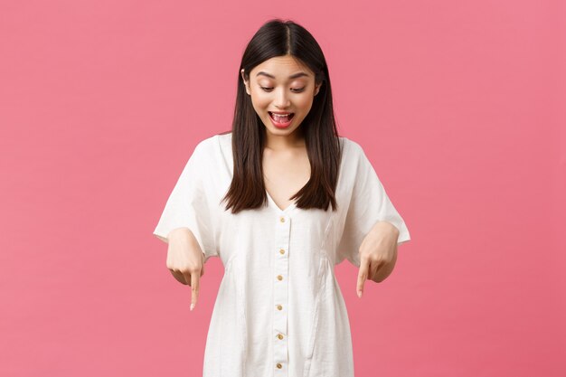 Belleza, emociones de la gente y concepto de ocio y vacaciones de verano. Chica asiática kawaii sorprendida y emocionada en vestido blanco, apuntando y mirando hacia abajo con cara feliz divertida, fondo rosa