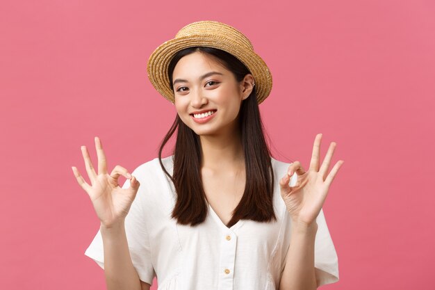 Belleza, emociones de la gente y concepto de ocio y vacaciones de verano. Sonriente niña asiática feliz con sombrero de paja que muestra un gesto bien, recomienda hotel o centro turístico perfecto, de pie con fondo rosa.