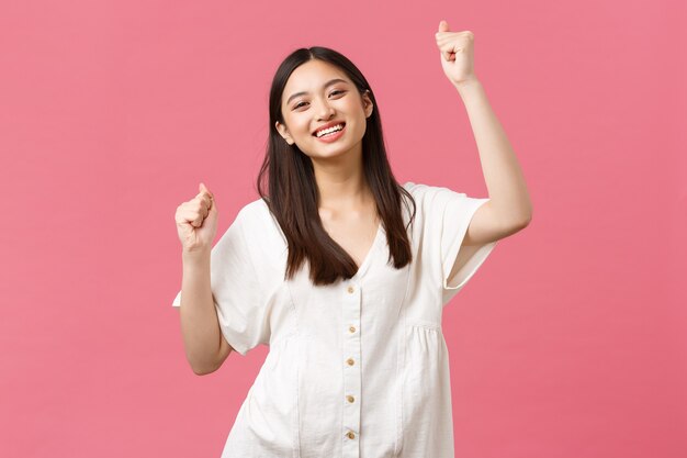 Belleza, emociones de la gente y concepto de ocio de verano. Alegre niña asiática carismática cantando, sintiendo felicidad y alegría, celebrando en la fiesta, bailando animadamente con las manos en alto, sonriendo