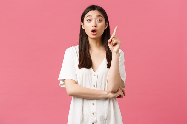 Belleza, emociones de la gente y concepto de ocio de verano. Emocionada chica asiática guapa con vestido blanco tiene una idea, levanta el dedo índice y sugiere una solución, cuenta su plan, fondo rosa.