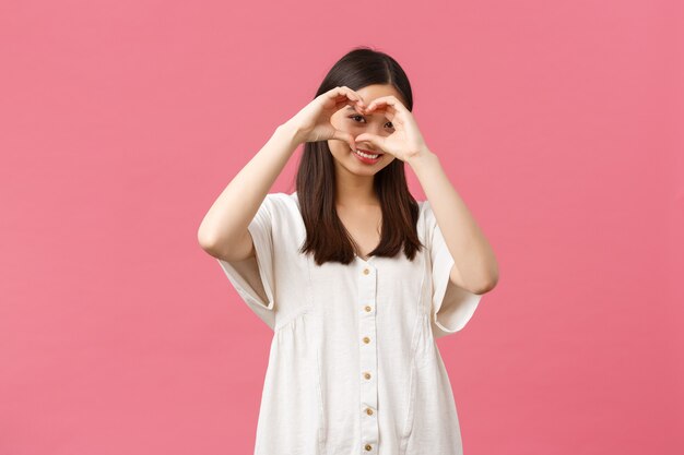 Belleza, emociones de la gente y concepto de ocio de verano. Encantadora y romántica chica asiática tímida en vestido blanco confiesa simpatía o amor, como alguien, mostrando el signo del corazón y sonriendo sobre fondo rosa.