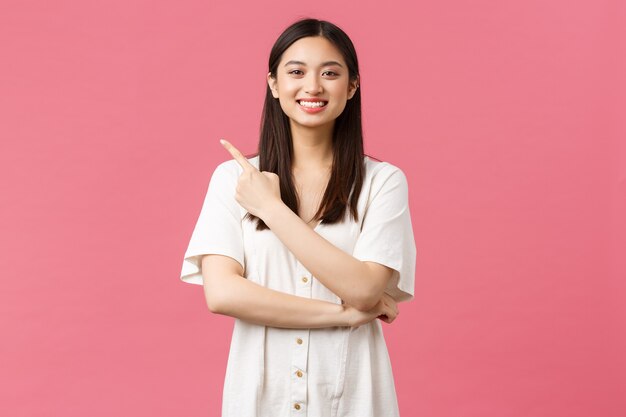 Belleza, emociones de la gente y concepto de ocio de verano. Hermosa mujer asiática entusiasta en vestido sonriendo a la cámara mientras señala el anuncio de la esquina superior izquierda sobre fondo rosa.