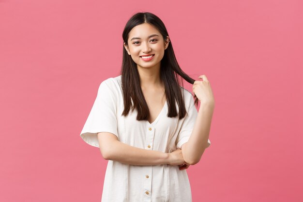 Belleza, emociones de la gente y concepto de ocio de verano. Linda novia coreana despreocupada con vestido blanco, sonriendo y tocando el mechón de cabello con una mirada feliz y soñadora, fondo rosa de pie