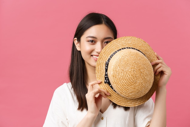 Belleza, emociones de la gente y concepto de ocio de verano. Primer plano de la cara de la cubierta de la chica japonesa tímida y linda detrás del sombrero de paja y sonriente fondo romántico rosa sensual, de pie.