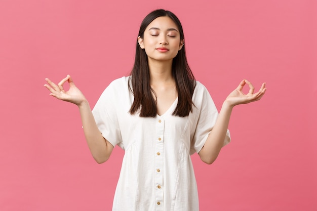 Belleza, emociones de la gente y concepto de ocio de verano. Relajada y tranquila, decidida joven asiática meditando en vestido blanco, cerrar los ojos y hacer ejercicios de respiración de yoga, fondo rosa.