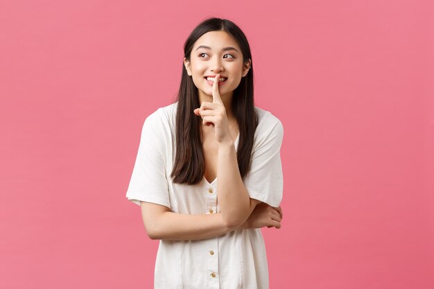 Belleza, emociones de la gente y concepto de ocio de verano. Soñadora y linda, encantadora chica asiática callando, pidiendo callarse o no contarle a nadie su secreto, apartando la mirada y sonriendo mientras cotillea.