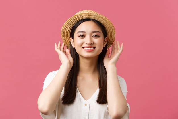 Belleza, emociones de personas y concepto de ocio y vacaciones. Encantadora mujer asiática de compras en la tienda, recogiendo un nuevo sombrero de paja, sonriendo encantada, comprando ropa de verano sobre fondo rosa