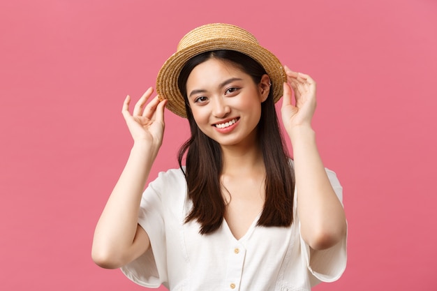 Belleza, emociones de personas y concepto de ocio y vacaciones. Encantadora mujer asiática de compras en la tienda, recogiendo un nuevo sombrero de paja, sonriendo encantada, comprando ropa de verano sobre fondo rosa.