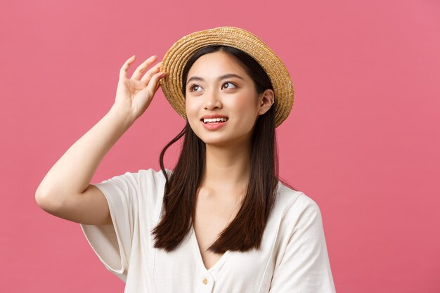 Belleza, emociones de personas y concepto de ocio y vacaciones. Encantadora mujer asiática de compras en la tienda, recogiendo un nuevo sombrero de paja, sonriendo encantada, comprando ropa de verano sobre fondo rosa.