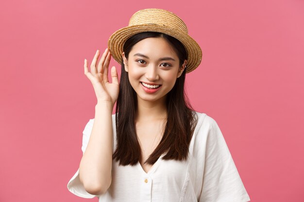 Belleza, emociones de personas y concepto de ocio y vacaciones. Encantadora mujer asiática de compras en la tienda, recogiendo un nuevo sombrero de paja, sonriendo encantada, comprando ropa de verano sobre fondo rosa.